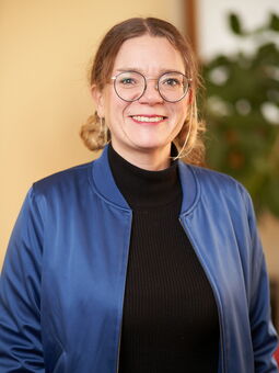 Porträt von Annika Harder. Sie hat honigblondes Haar und einen Mittelscheitel. Das Haar hat sie zu zwei Zöpfen zusammengebunden. Außerdem trägt sie eine schwarzgeränderte Metallbrille, einen schwarzen Pullover und darüber eine azurblaue Jacke