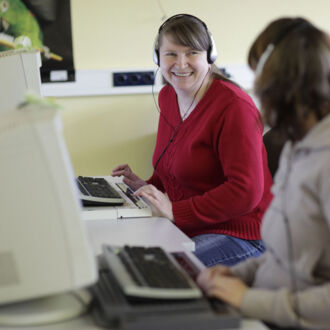 Zwei Frauen arbeiten an Computern, die mit Blindenhilfstechnik ausgestattet ist. Dabei unterhalten sie sich