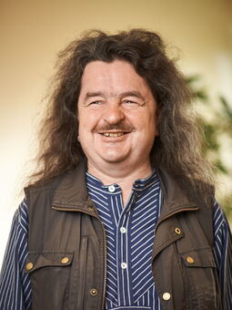 Robbie Sandberg trägt sein braunes gewelltes Haar schulterlang und einen Oberlippenbart. Er lächelt freundlich in die Kamera. Gekleidet ist er mit einem Fischerhemd, darüber eine braune Weste