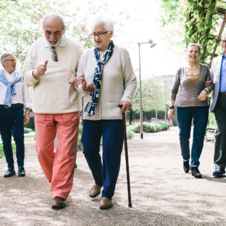 Eine Gruppe Senioren und Seniorinnen geht paarweise mit Spazierstöcken und miteinander untergeharkt spazieren.