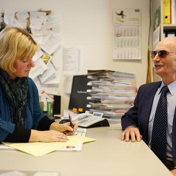 EUTB-Beraterin Annette Schacht füllt ein Formular für einen ratsuchenden Herrn aus. Sie sitzen an ihrem Schreibtisch