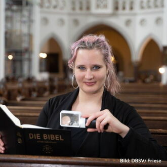 Eine Frau Mitte 20 sitzt in einer Kirchenbank und lächelt in die Kamera, in einer Hand eine Leuchtlupe, in der anderen eine Bibel. Sie hat langes, dunkelblondes Haar mit rötlichen Strähnchen und trägt große Ohrringe.