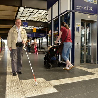 Ein blinder Mann läuft durch einen Bahnhof. Mit seinem Langstock orientiert er sich am Leitsystem auf dem Boden