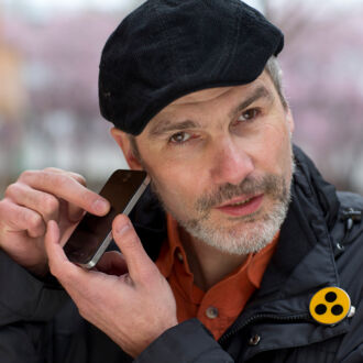 Ein Mann bedient sein Smartphone und hält es dabei ans Ohr. Eine Plakete am Revers kennzeichnet ihn als blinden Menschen.
