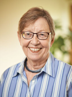 Brigitte Kukies hat honigfarbenes, kurzgeschnittenes Haar und trägt eine dunkel geränderte, eckige Brille. Gekleidet ist sie mit einer blau-weiß gestreiften Bluse und einer blauen Halskette.