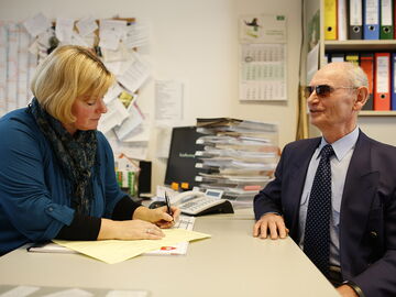EUTB-Beraterin Annette Schacht füllt ein Formular für einen ratsuchenden Herrn aus. Sie sitzen an ihrem Schreibtisch