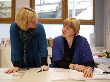 Die EUTB-Beraterinnen Annette Schacht und Christin Becker beraten sich. Frau Becker sitzt am Schreibtisch, Frau Schacht steht neben ihr und beugt sich herunter