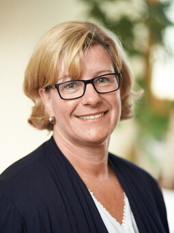 Annette Schacht hat blondes Haar bis zu den Ohren mit einem seitlichem Pony. Sie lächelt in die Kamera und trägt ein weißes Oberteil und eine dunkelblaue Strickjacke.