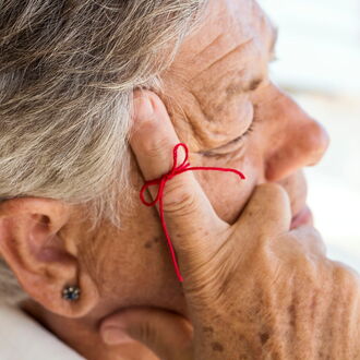 Nahaufnahme einer älteren Frau im Profil. In nachdenklicher Pose hält sie sich die Hand an ihr Gesicht, den Zeigefinger an der Schläfe. Am Zeigefinger ist eine Schleife mit einem roten Faden gebunden, als solle er sie an etwas erinnern.