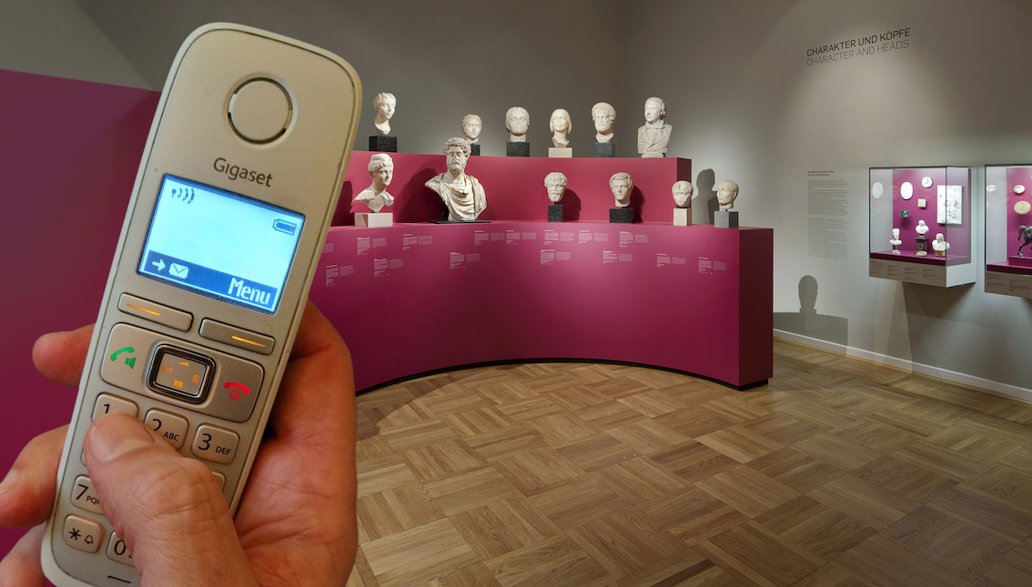 Im Bildhintergrund sieht man auf einer Gallerie verschiedene Büsten. Im Vordergrund hält eine Hand ein Telefon