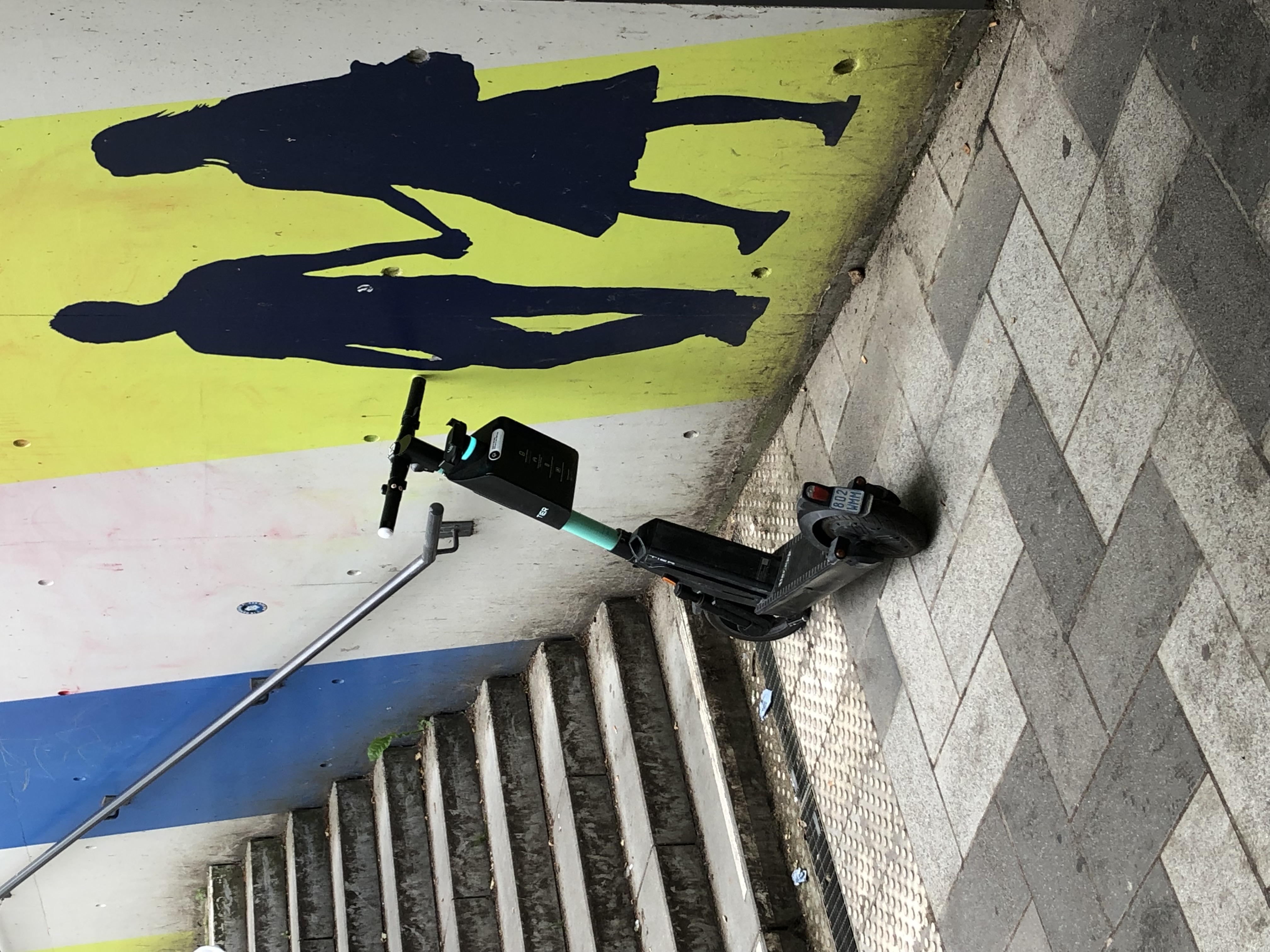 Ein E-Roller in einer Bahnstation vor dem Treppenaufgang. Er versperrt den Weg zum Handlauf an der Treppe